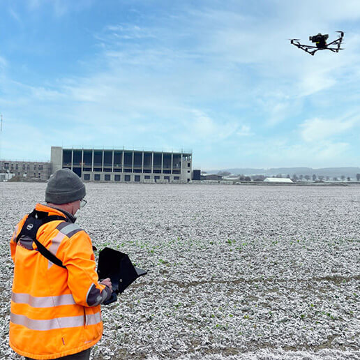 Person in Sicherheitskleidung bedient eine Drohne in einem kahlen Feld vor einem modernen Gebäude