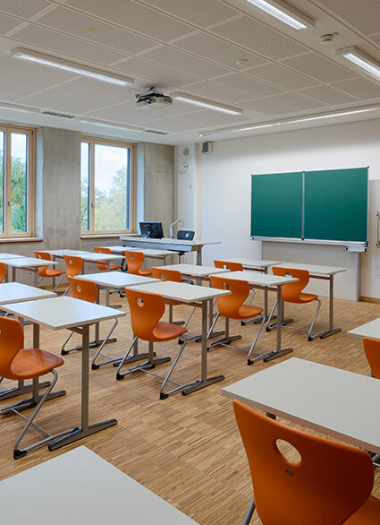 Klassenzimmer der FOS/BOS Krumbach mit Reihen von Tischen und Stühlen, einer grünen Tafel an der Wand und Fenstern, die auf Grünanlagen blicken, beleuchtet durch Deckenleuchten